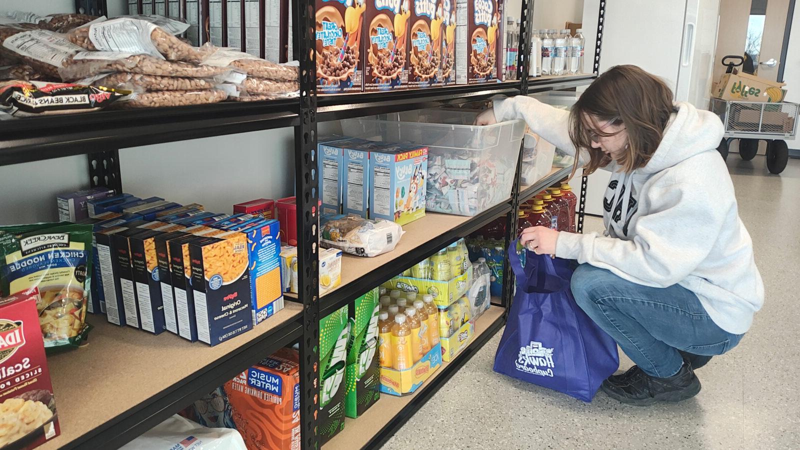 A woman wearing a gray hoodie and blue jeans is crouching down while selecting items from a shelf stocked with food and drinks. They are placing the items into a blue reusable bag with the logo "Hawk Cupboard." The shelves contain various goods, including cereal boxes, pasta, canned goods, and bottled beverages.